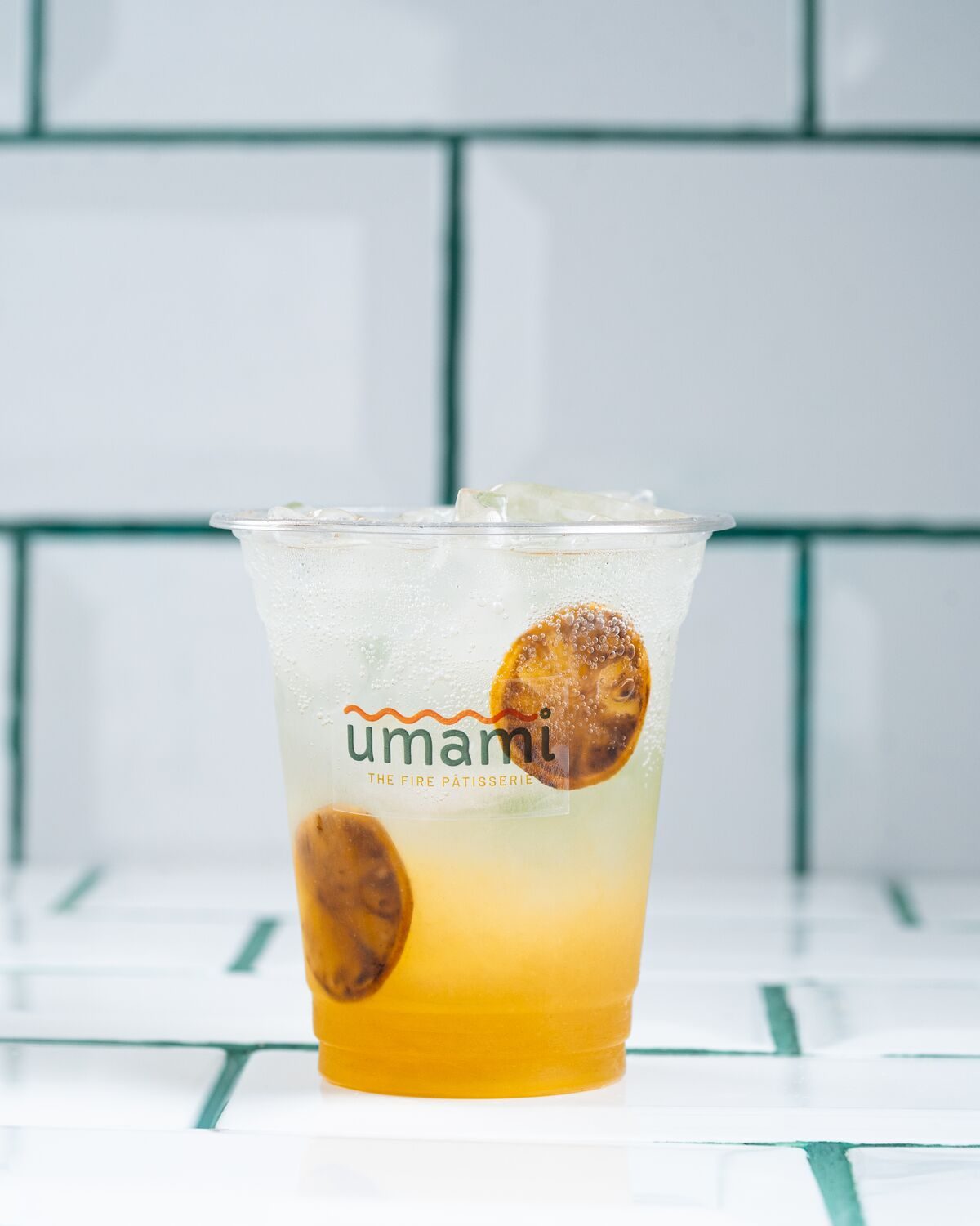 A clear cup of layered yellow and amber charred lemonade over ice, garnished with two blackened lemon slices.