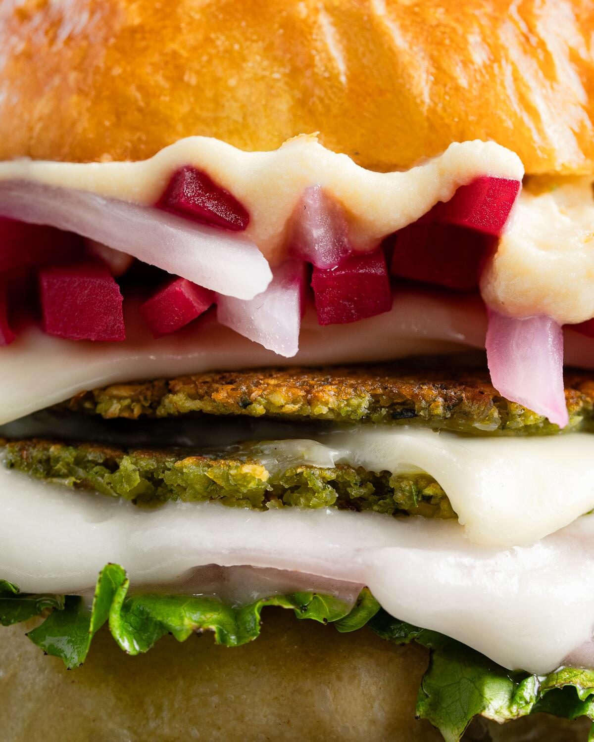 Close up of two green herbed falafel patties with melted white cheese, diced beetroot, sliced onion and frilled lettuce in a soft milk bun.