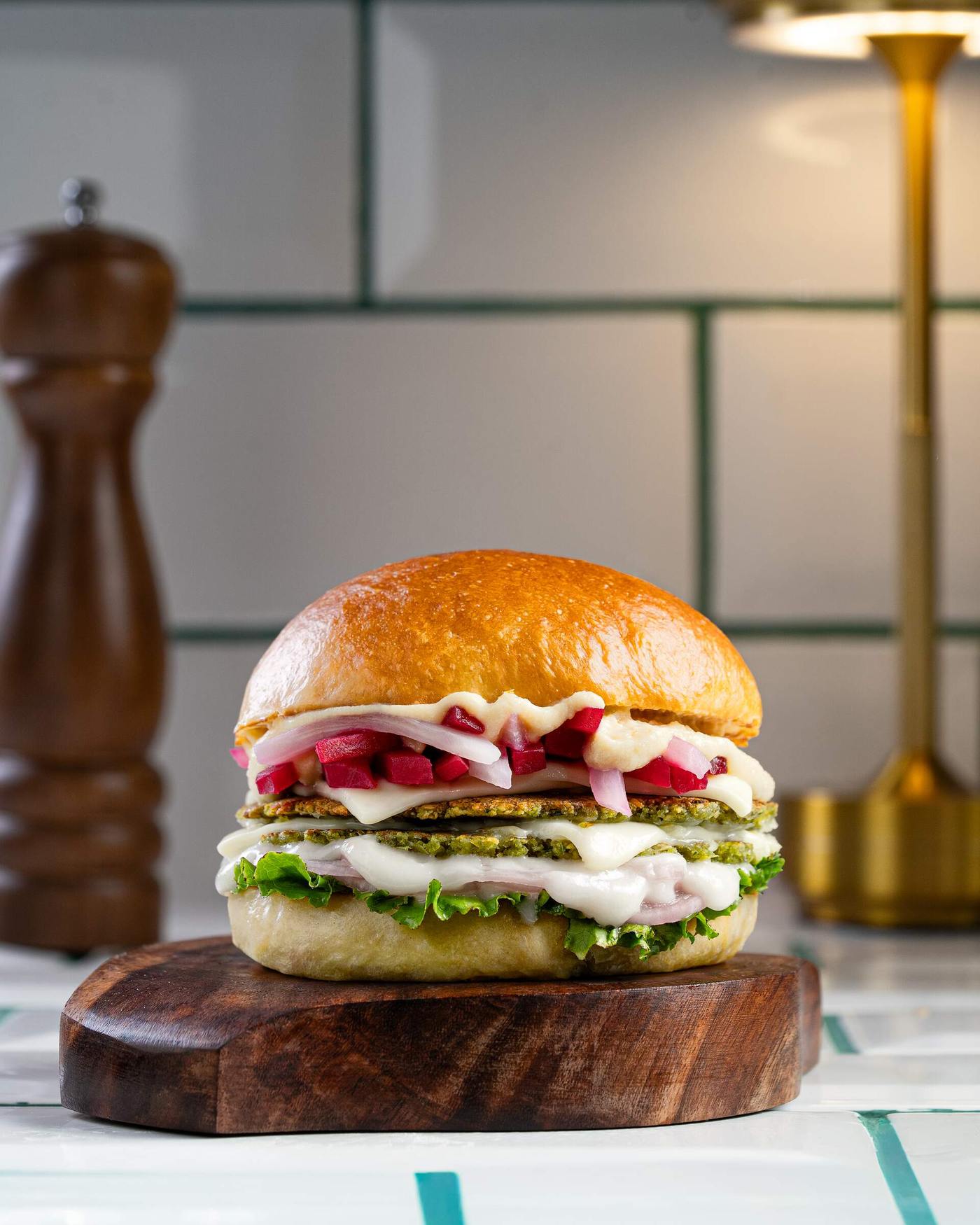 Two green herbed falafel patties layered with melted white cheese, diced beetroot, sliced onion and lettuce in a soft milk bun on a dark wood board.