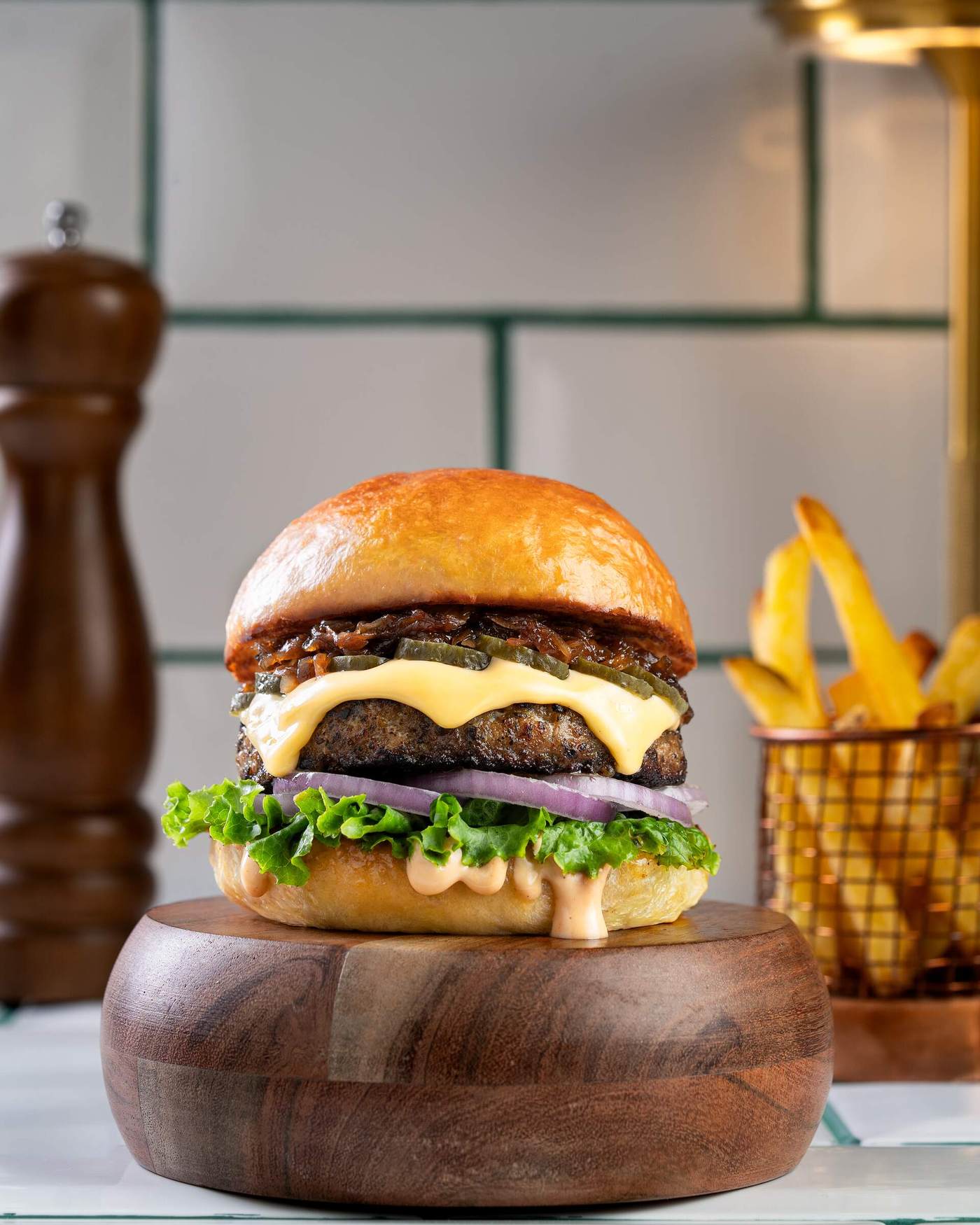 A thick fire-grilled chicken patty topped with melted cheese, sliced pickles and caramelised onion, stacked with red onion and lettuce in a glossy milk bun on a dark wood board.
