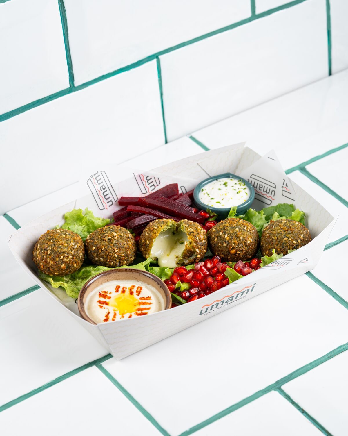 Green herbed falafel balls in a paper tray with lettuce, sliced beetroot, pomegranate seeds and two creamy dips, shot at a forty five degree angle.