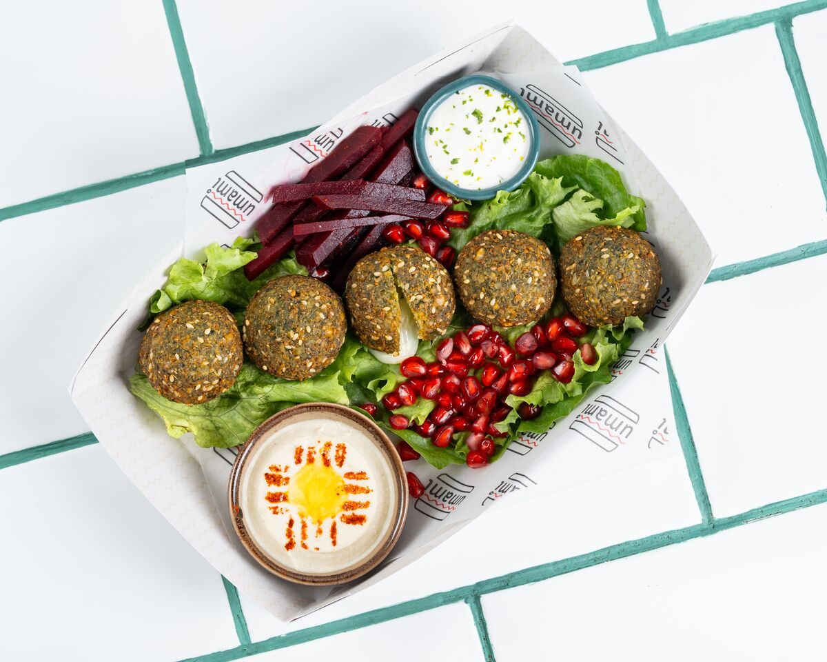 Green herbed falafel balls in a paper tray with lettuce, sliced beetroot, pomegranate seeds and two creamy dips, shot from directly overhead.