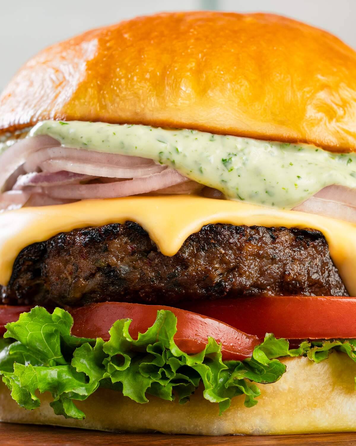 Close up of a thick char-grilled mutton patty under melted cheese, layered with sliced red onion, herbed green mayonnaise, a tomato slice and frilled lettuce.