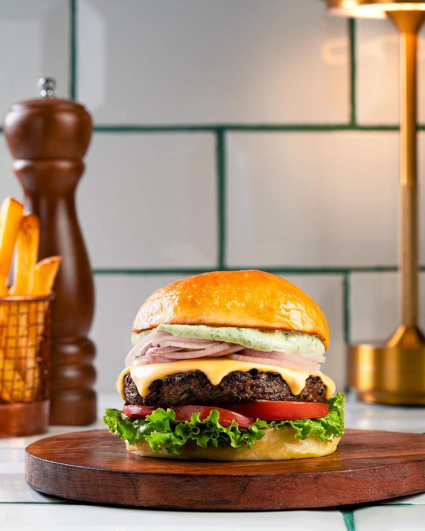 A thick char-grilled mutton patty with melted cheese, sliced red onion, herbed green mayonnaise, tomato and lettuce in a glossy milk bun on a dark wood board.